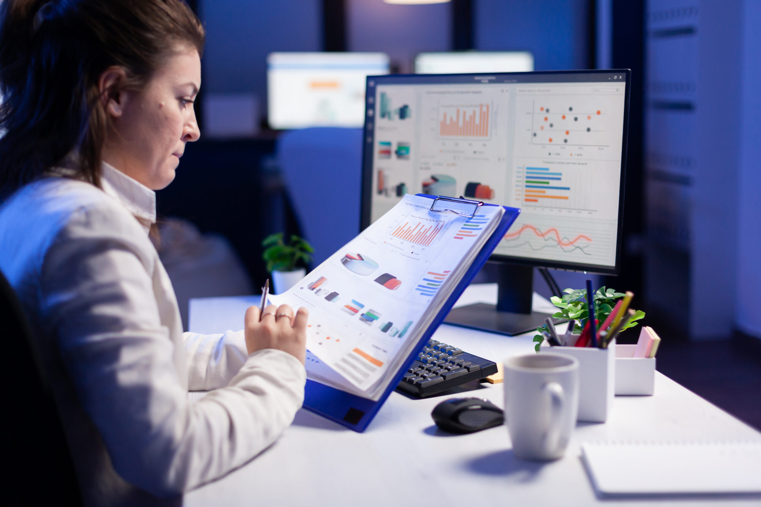 Freelancer woman comparing graphics from clipboard with grafics from computer at business office. Tired focused employee using modern technology network wireless working on project solution